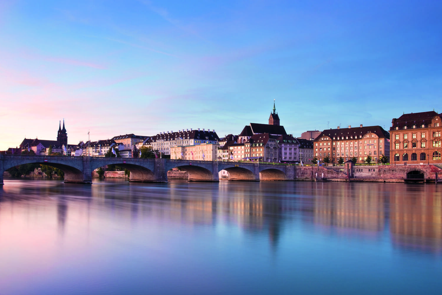 Steinbrücke vor historischen Gebäuden am ruhigen Fluss bei Abendlicht.