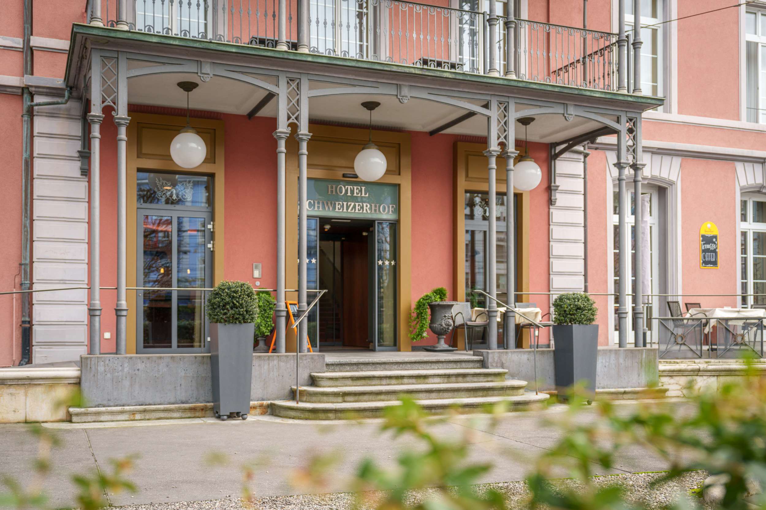 Eingang eines Hotels mit überdachter Terrasse, Treppe und Pflanzkübeln vor einer rosafarbenen Fassade.